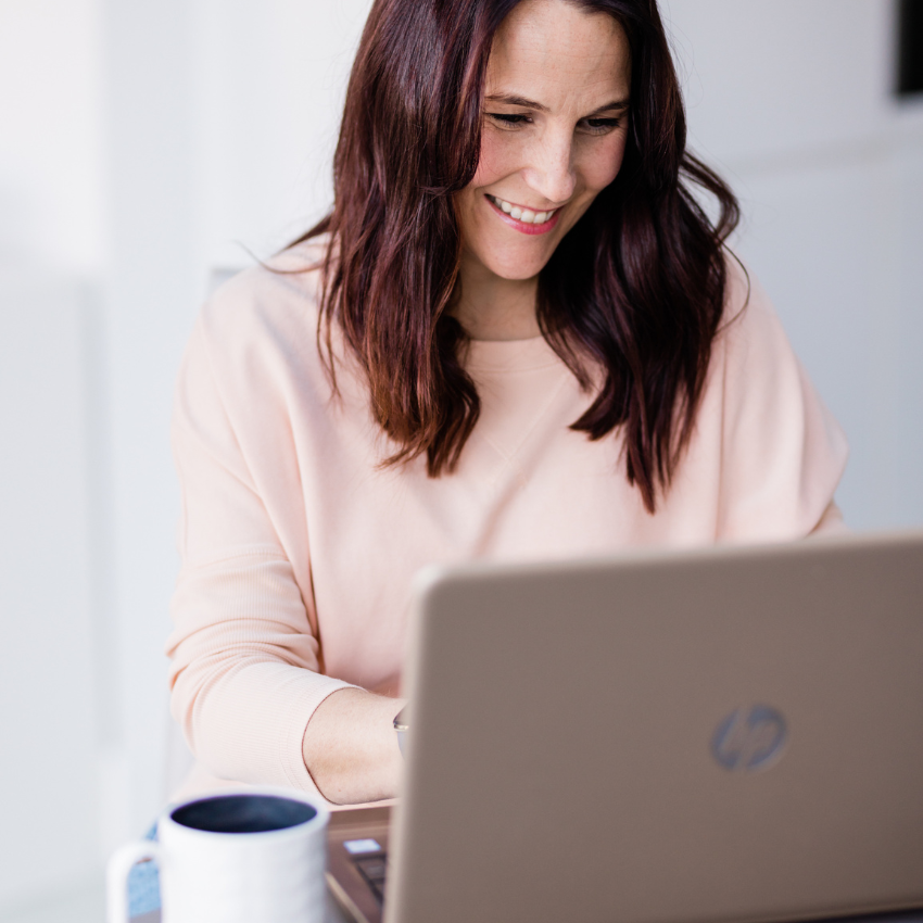 Dark haired, Caucasian woman in a peach jumper sitting at a laptop