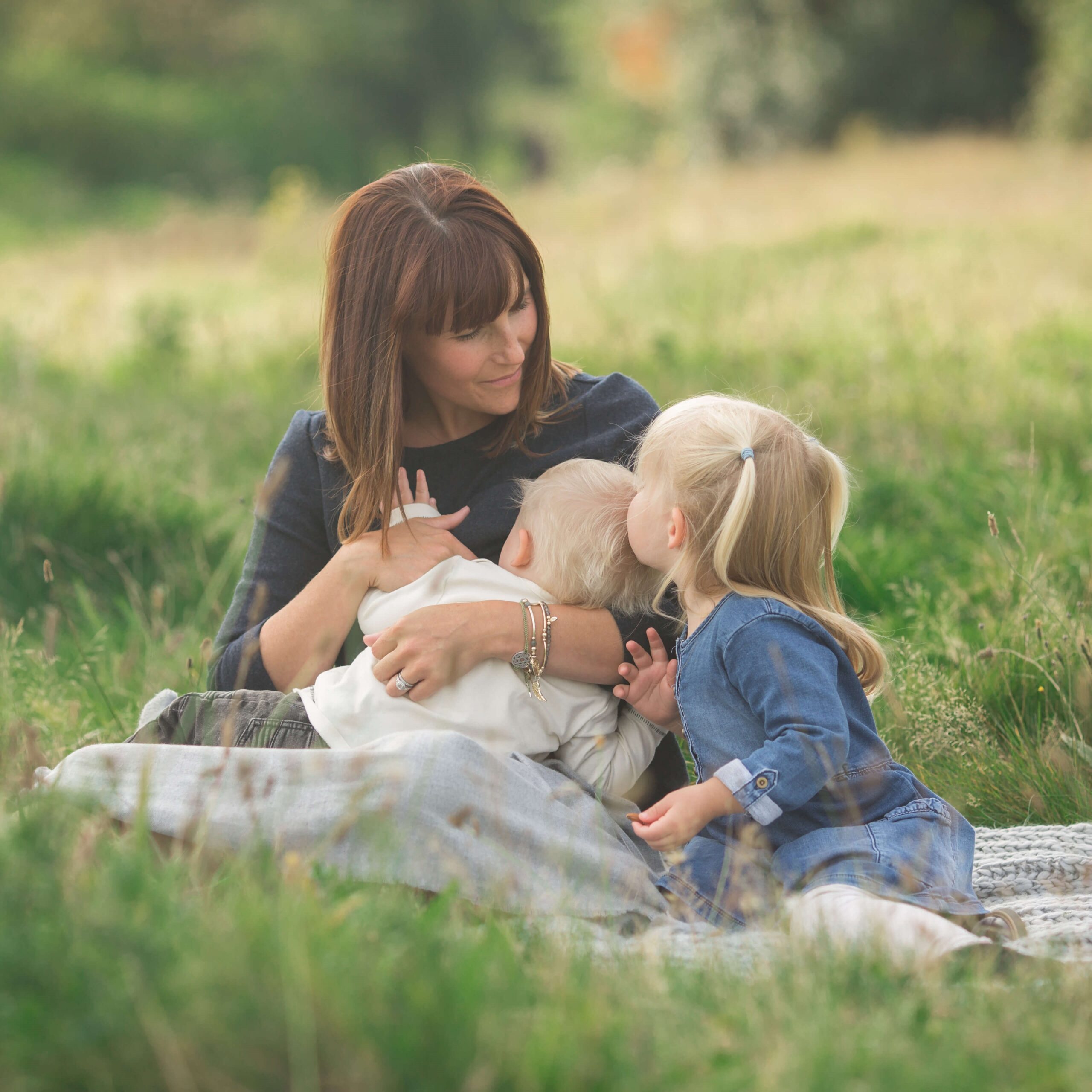 Dark haired women sitting in long grass, breastfeeding blonde baby. Blonde female toddler kissing baby on the head